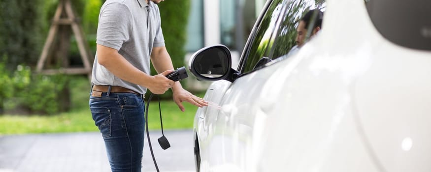 Progressive asian man install cable plug to his electric car with home charging station in the backyard. Concept use of electric vehicles in a progressive lifestyle contributes to clean environment.