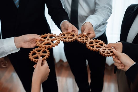 Closeup hand holding wooden gear by businesspeople wearing suit for harmony synergy in office workplace concept. Group of people hand making chain of gears into collective form for unity symbol.