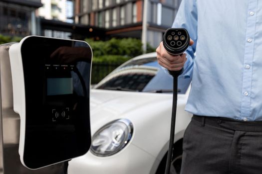 Closeup progressive hand holding EV charger plug and pointing at camera with his electric car recharging at charging station near city residential apartment condo building in the background.