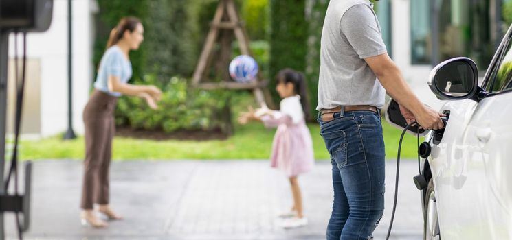 Focus image of progressive man charging electric car from home charging station with blur mother and daughter playing together in the background.