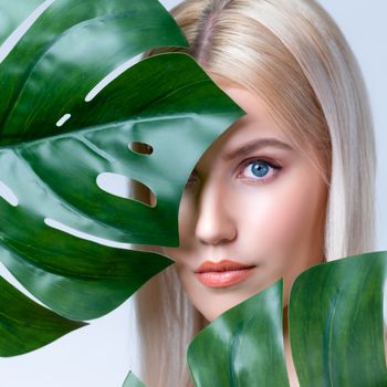 Closeup facial portrait personable woman with perfect smooth makeup holding green monstera leaves and cover her face as natural healthy skincare treatment. Tropical nature and beauty concept