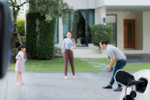 Focus closeup electric vehicle recharging battery from home electric charging station with blurred family in background. Renewable clean energy car for progressive eco awareness lifestyle concept.