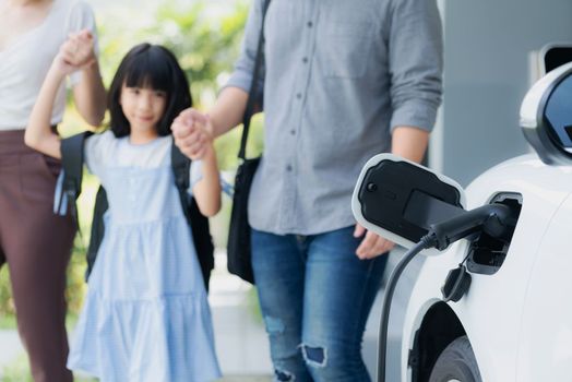 Focus closeup electric vehicle recharging battery from home electric charging station with blurred family in background. Renewable clean energy car for progressive eco awareness lifestyle concept.