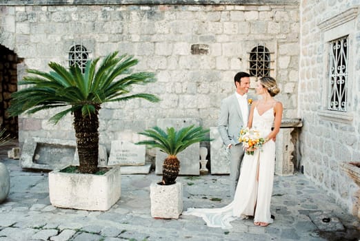 Bride with a bouquet stands next to groom in the courtyard of an old stone building. High quality photo