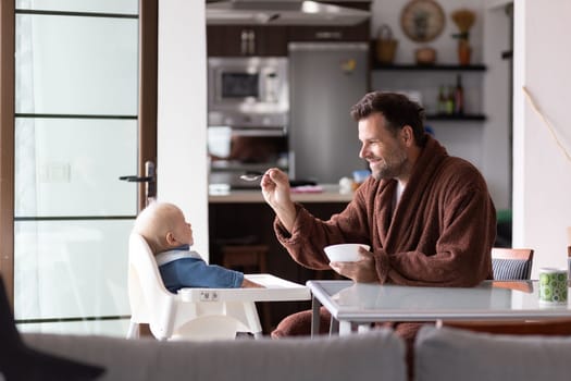 Father wearing bathrope spoon feeding hir infant baby boy child sitting in high chair at the dining table in kitchen at home in the morning