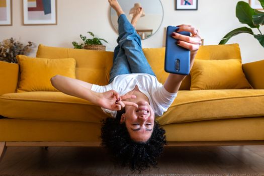 Joyful African American woman lying upside down on the sofa takes selfie with smart phone. Social media concept.