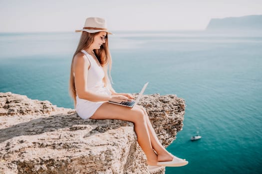 Successful business woman in yellow hat working on laptop by the sea. Pretty lady typing on computer at summer day outdoors. Freelance, travel and holidays concept.