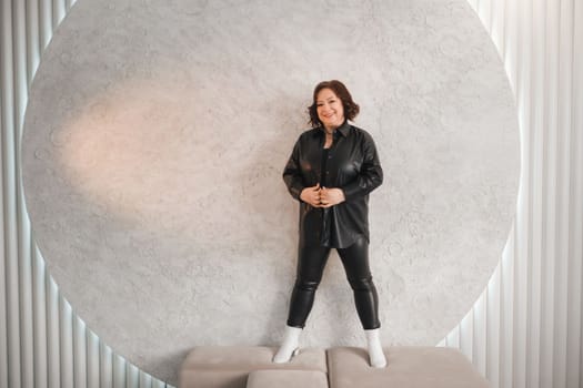 An adult woman in black leather clothes poses against a gray studio wall.