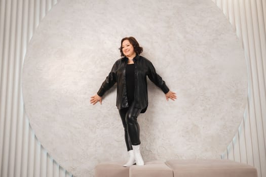 An adult woman in black leather clothes poses against a gray studio wall.