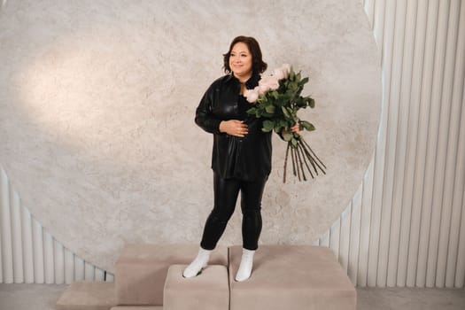 a stylish adult woman in black leather clothes stands with a bouquet of pink roses in the interior.