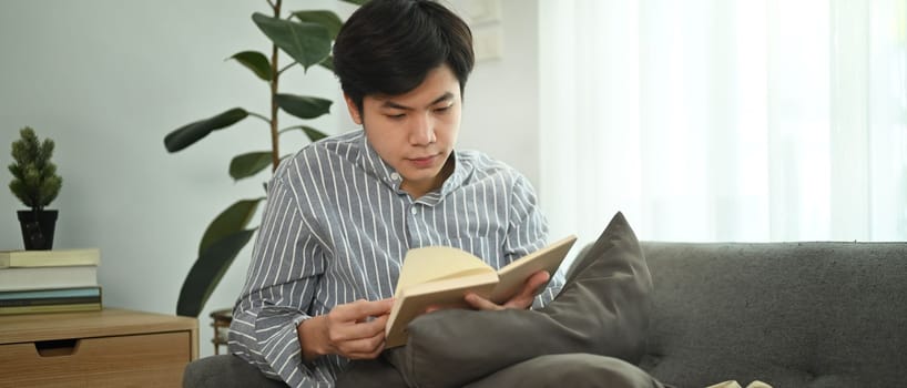 Calm asian man sitting on a comfortable couch and reading book, spending leisure weekend time at home.