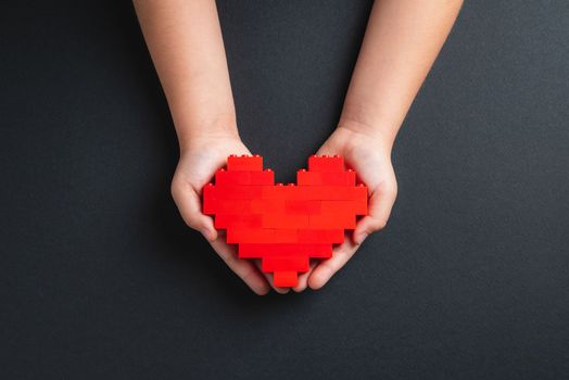 Hands of little girl child holding heart made of plastic bricks on dark gray background