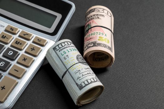 Rolls of 50 and 100 us dollar standing near calculator on dark background