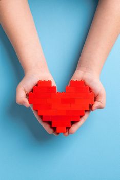 Hands of little girl child holding heart made of plastic bricks on blue background