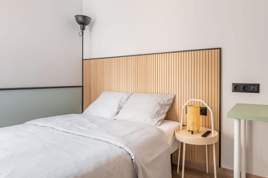 Part of a bedroom with a double bed against a white wall with designer wooden elements. Floor lamp with black metal shade in the corner. On the bedside table is a night lamp with a yellow shade