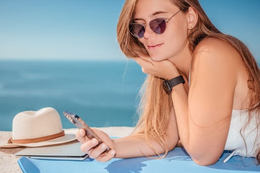 Successful business woman in yellow hat working on laptop by the sea. Pretty lady typing on computer at summer day outdoors. Freelance, travel and holidays concept.