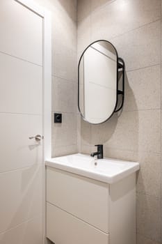 Corner of a bathroom with a white sink on a vanity with two large drawers for bathroom amenities. Above the sink is an oval designer mirror. Decorative elements made of black metal