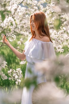 Woman cherry orchard. A happy woman in a long white long dress walks through the green spring blooming cherry garden. Happy cheerful princess bride. The fabric of the skirt flutters in the wind