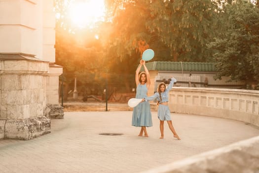 Daughter mother run holding hands. In blue dresses with flowing long hair, they hold balloons in their hands against the backdrop of a sunset and a white building