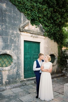 Groom hugs bride from behind near the weathered building in the garden. High quality photo