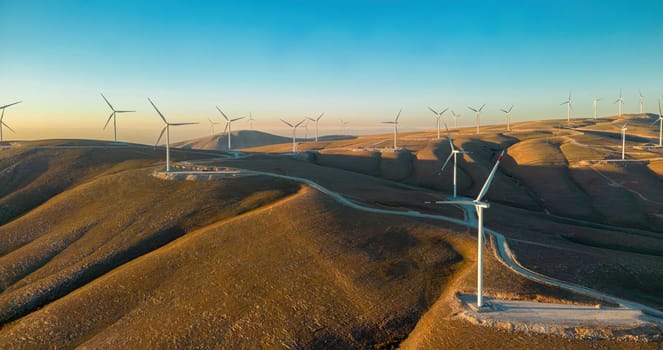 Aerial view of multiple wind turbines standing on a hill and generating electricity at sunrise