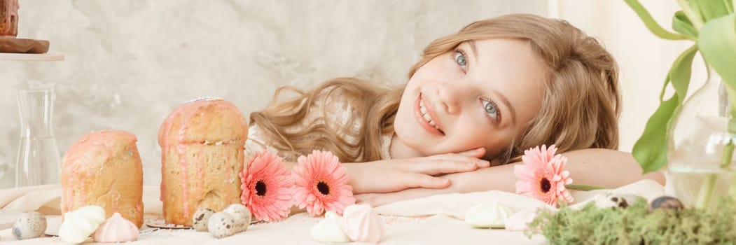 A girl with long hair in a light dress is sitting at the Easter table with cakes, spring flowers and quail eggs. Happy Easter celebration