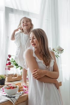 A little blonde girl with her mom on a kitchen countertop decorated with peonies. The concept of the relationship between mother and daughter. Spring atmosphere.