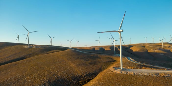 Aerial view of multiple wind turbines standing on a hill and generating electricity at sunrise