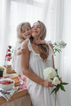 A little blonde girl with her mom on a kitchen countertop decorated with peonies. The concept of the relationship between mother and daughter. Spring atmosphere.