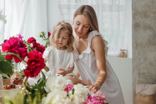 A little blonde girl with her mom on a kitchen countertop decorated with peonies. The concept of the relationship between mother and daughter. Spring atmosphere.