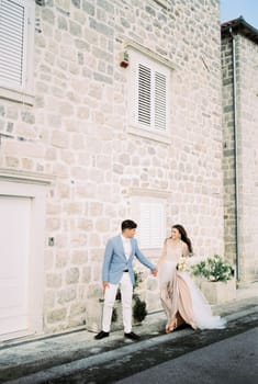 Bride and groom walk hand in hand past an old stone building. High quality photo