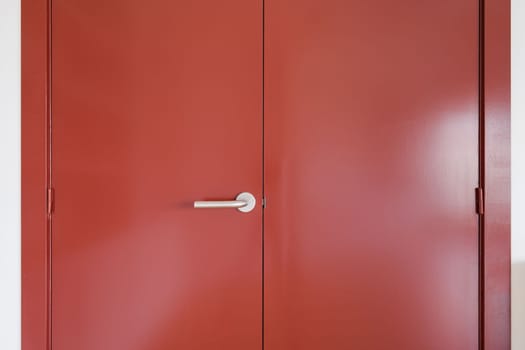 Closeup of a wide front double-leaf wooden door covered with red paint. Door handle made of durable metal. The door is held on door hinges. Sunlight sets off the depth of color