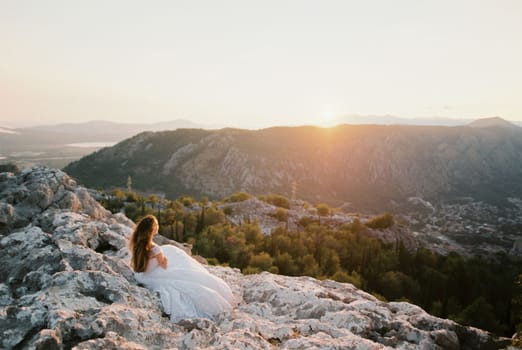Bride in a white dress sits on a rock and looks at the sunset over the mountains. High quality photo