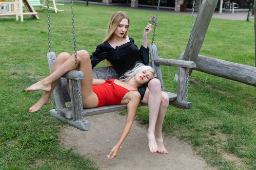 Intimate relationship. Two young women on wooden swings outdoors. Blonde woman in red lies on brunette woman's lap in black dress holding a bouquet of flowers.