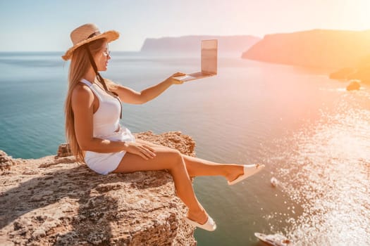 Working remotely on seashore. Happy successful woman female freelancer in straw hat working on laptop by the sea at sunset. Freelance, remote work on vacation