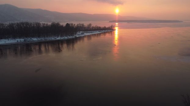 View of the hills by the river at sunset. Orange sun over my river. Snow lies on the island. Top view from a drone. 4k