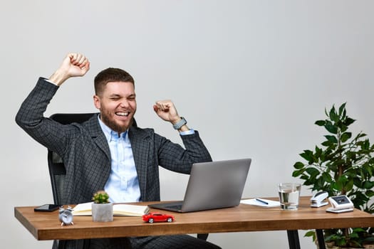Satisfied young man gesturing emotionally and celebrating victory while sitting on chair at desk, using laptop pc computer