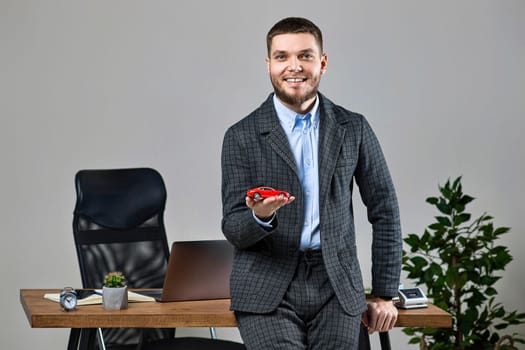 Successful caucasian cheerful man holding a toy car while standing at desk. Car dealerships offer car . Car insurance