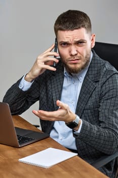 young bearded businessman sitting on chair at desk and talking on the phone, using laptop