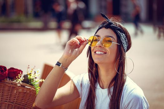 Portrait of a happy smiling young woman who is enjoying in a summer sunny day, sitting on a city bench and looking through sunglasses, beside the bike with flower basket.