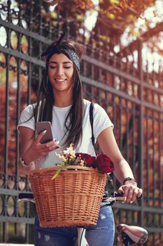 Smiling young woman reading message on smartphone on the city street, on a sunny day, beside the bike with flower basket.