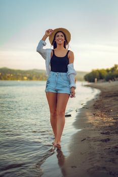 Young smiling woman in straw hat relaxing at sunset time on the river bank. She is walking along river beach and smiling.