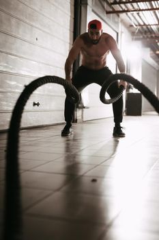 Handsome muscular young man exercising with ropes at the garage gym. Selective focus.