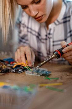 Young woman technician focused on the repair of electronic equipment by soldering iron.
