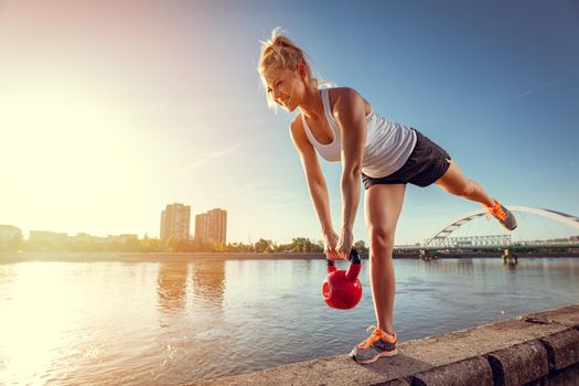Young fitness woman doing workout with kettlebell on the wall  by the river in a sunset.
