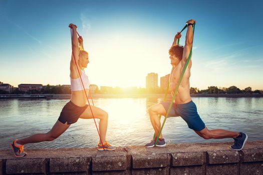 Young fitness couple doing workout with rubber band by the river in a sunset. 