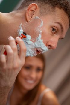 Portrait of a beautiful young man is shaving in front of his bathroom mirror. Selective focus.