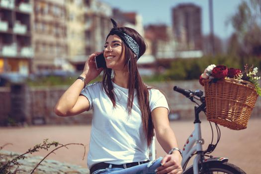 Laughing young woman talking on smartphone sitting on a little wall in a sunny day, beside the bike with flower basket.