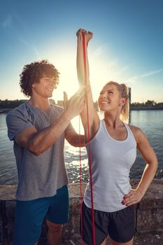 Young fitness couple doing workout with rubber band by the river in a sunset. The woman is stretching arms and the man support her.