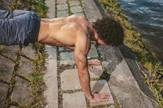 Focused muscular young sport man with naked torso, doing push-ups during outdoor cross training by the river.